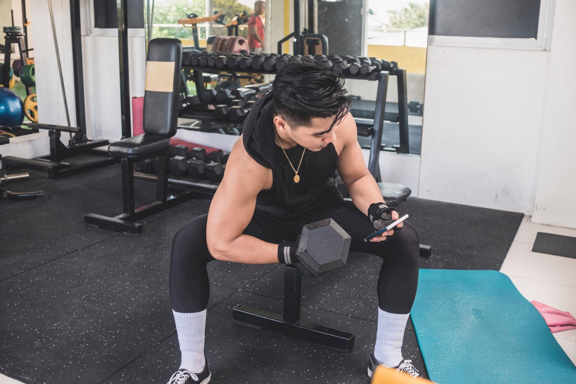 Man in gym checking workout plan on phone between sets with dumbbell